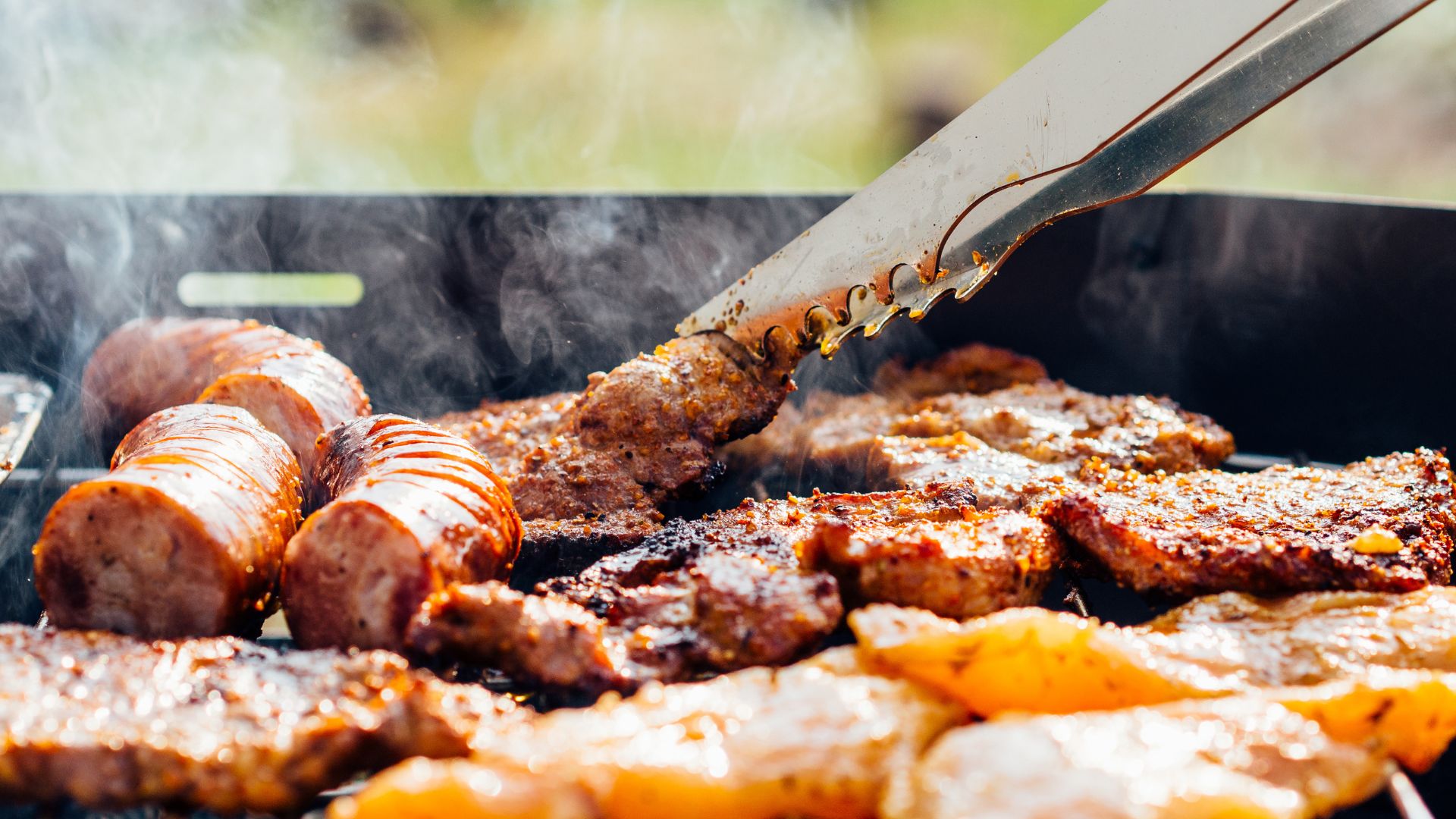Assorted meats grilling on a barbecue with tongs